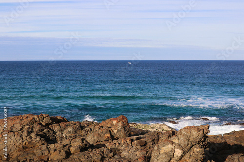 Humpback whale swimming in the distance with it's tail out of the water off of rocks. Waves crashing on the rocks with the beautiful blue and green ocean. Whale breaching on a summer day with blue sky