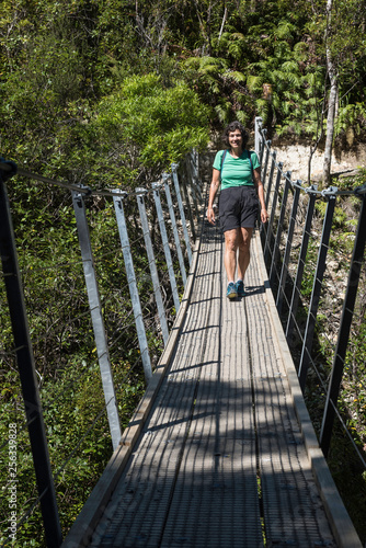 Looking along the wooden deck of a swing bridge at a female hiker crossing the Kauaeranga river in the Coromandel, New Zealand.