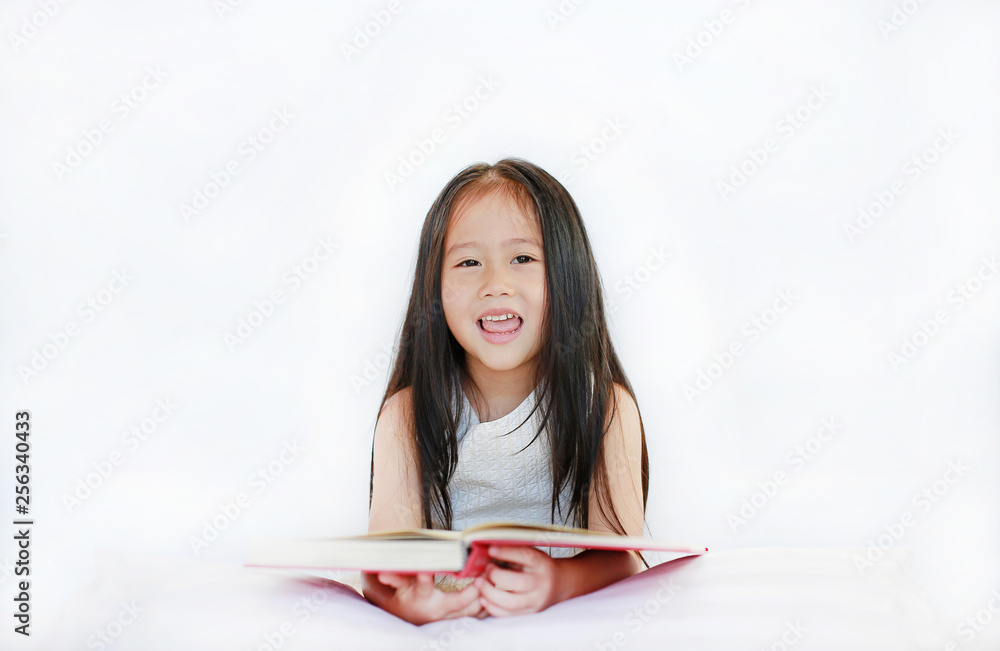 Smiling little Asian kid girl reading hardcover book lying with pillow on bed against white background. Smiling little Asian kid girl reading hardcover book lying with pillow on bed against white background.