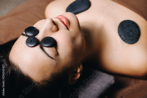 Close up portrait of a young european female having hot stone therapy on her face and shoulders while leaning on a spa bed with closed eyes in a wellness center.