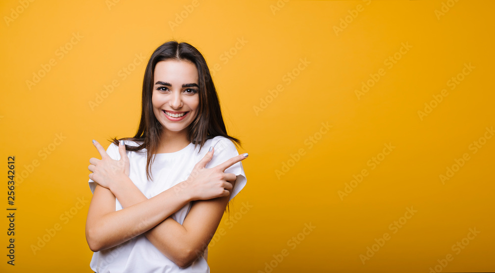 Obraz premium Beautiful young attractive girl with long dark hair looking at camera laughing while embracing herself isolated over yellow background.