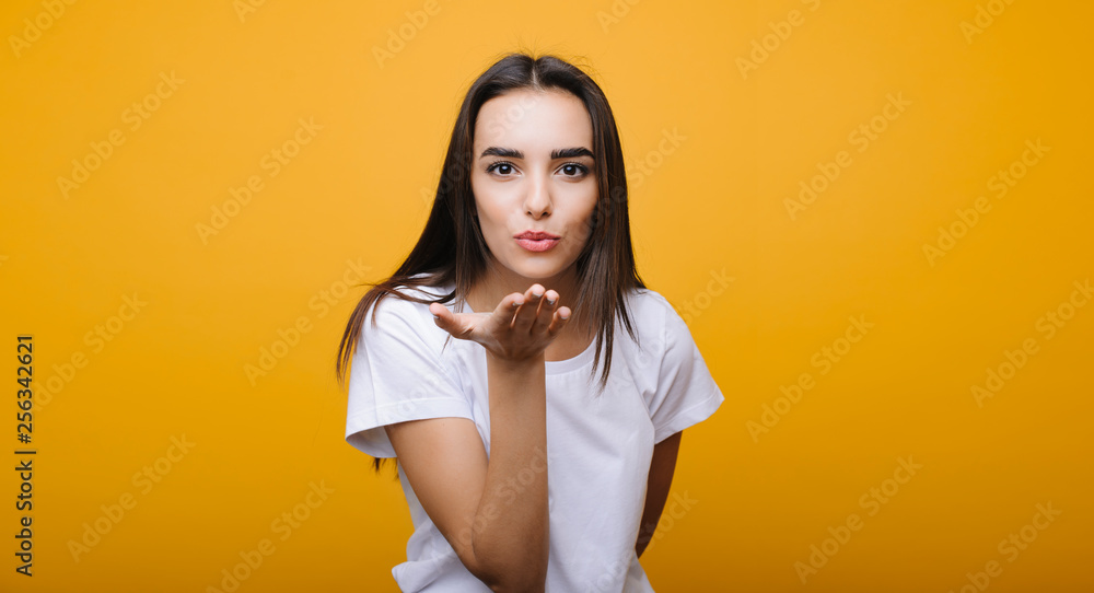 Obraz premium Portrait of a cute european female looking at camera and doing a air kiss while dressed in white shirt in front of a yellow studio background.