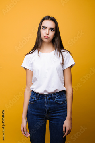 Portrait of a charming female with dark hair dressed in white shirt and jeans looking at camera and blowing her cheeks against yellow background.