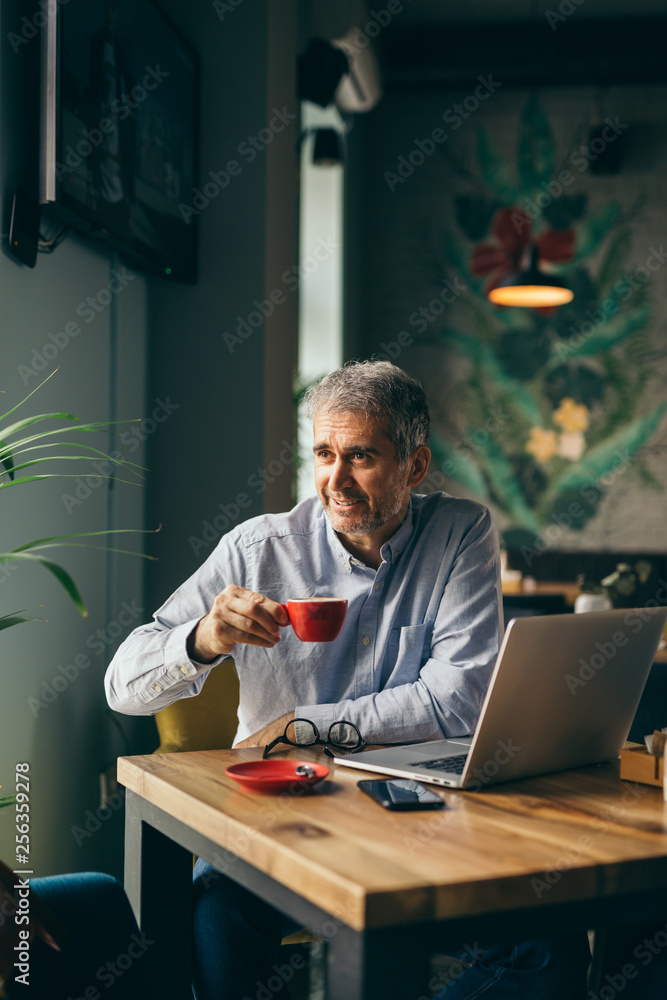 handsome gray haired man drinking coffee and working on laptop in cafeteria handsome gray haired man drinking coffee and working on laptop in cafeteria