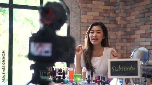 Pretty Asian woman recording video about cosmetics with DSLR on tripod with subscribe signboard. Main focus on camera screen