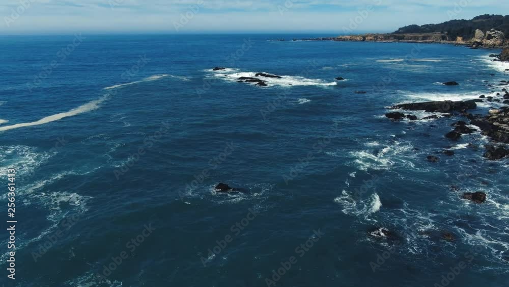 Sun drenched rocks are rhythmically beaten by Pacific Ocean Waves.  Cliffs and rugged trees frame the seashore.  Drone based aerial video. Cinematic and beautiful.  Both calming and energizing.