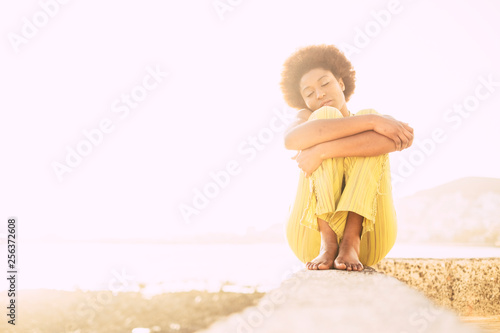 Lonely cute afro american hair style girl sitting and hugging herself in outdoor feeling the nature and sensations around - portrait of young black girl with clear white sky in background