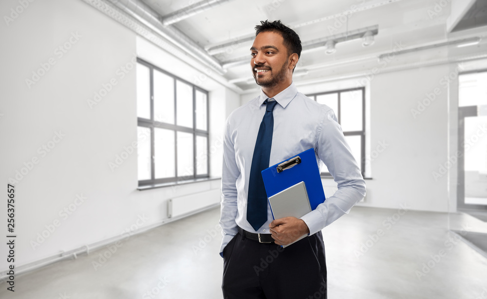 business, realty and people concept - smiling indian businessman or realtor with clipboard over empty office room background business, realty and people concept - smiling indian businessman or realtor with clipboard over empty office room background