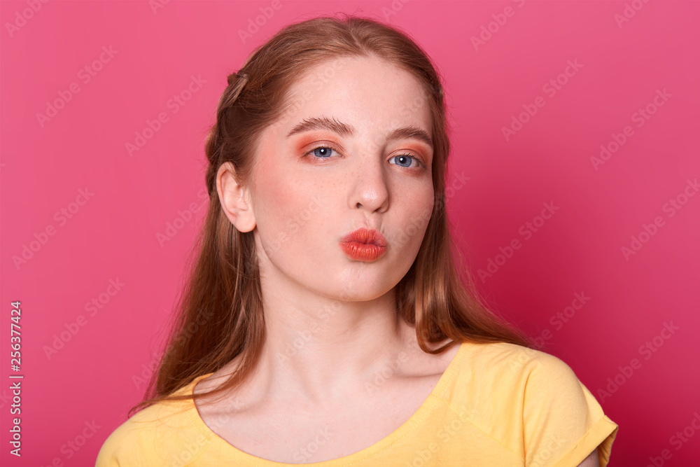 Portrait of lovely female with long straight hair, wears yellow t shirt, rounds lips as going to recieve kiss from boyfriend, has mysterious facial expression, isolated over pink background. Portrait of lovely female with long straight hair, wears yellow t shirt, rounds lips as going to recieve kiss from boyfriend, has mysterious facial expression, isolated over pink background.
