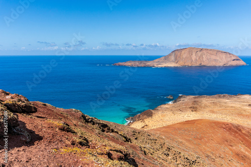Isla de Montana Clara island as seen from the top of Bermeja volcanic mountain on La Graciosa Island in Lanzarote, Spain. Colorful landscape with red rugged cliffs and turquoise sea waters.
