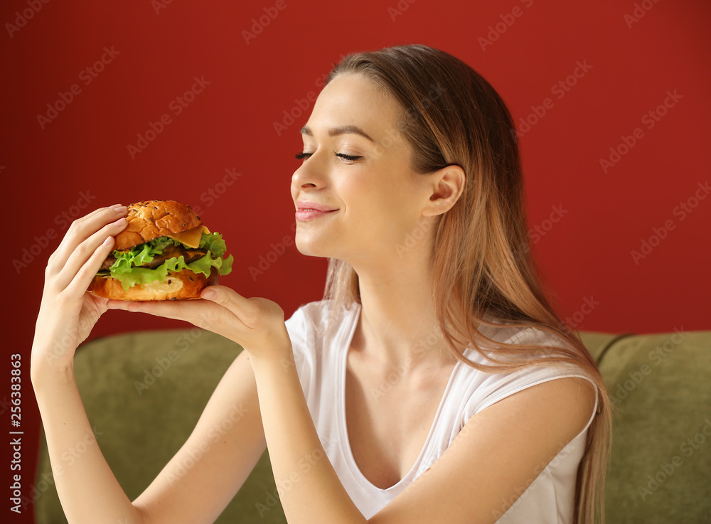 Beautiful young woman with tasty burger sitting on sofa Beautiful young woman with tasty burger sitting on sofa