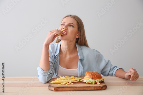 Young woman eating tasty burger with french fries at table