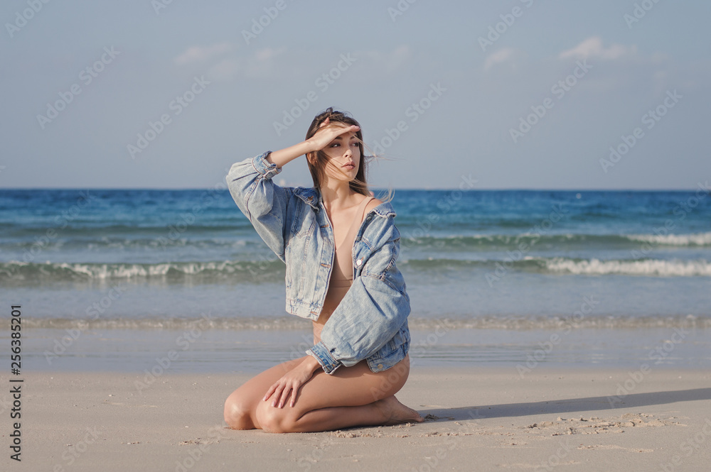 Young female model sitting on an empty beach by the ocean with waves and looking for something. Woman in beige casual swimwear and denim jacket Young female model sitting on an empty beach by the ocean with waves and looking for something. Woman in beige casual swimwear and denim jacket