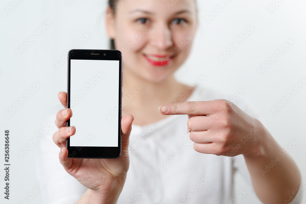 a young smiling woman holds a smartphone vertically with a white screen with a place for copyspace and points at him the finger of the other hand. on light background