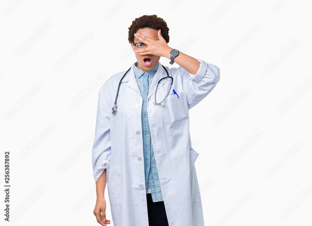 Young african american doctor woman wearing medical coat over isolated background peeking in shock covering face and eyes with hand, looking through fingers with embarrassed expression. Young african american doctor woman wearing medical coat over isolated background peeking in shock covering face and eyes with hand, looking through fingers with embarrassed expression.