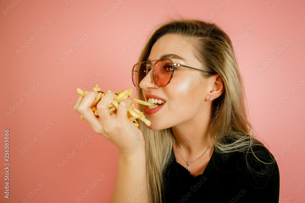 young beautiful blonde girl model with appetite eating fast food, French fries holding in hand, on a pink background young beautiful blonde girl model with appetite eating fast food, French fries holding in hand, on a pink background