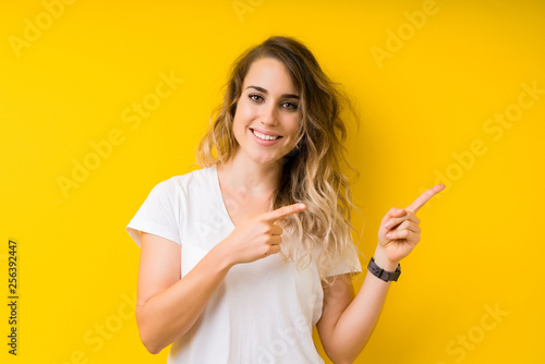 Young beautiful blonde woman over yellow background smiling and looking at the camera pointing with two hands and fingers to the side.