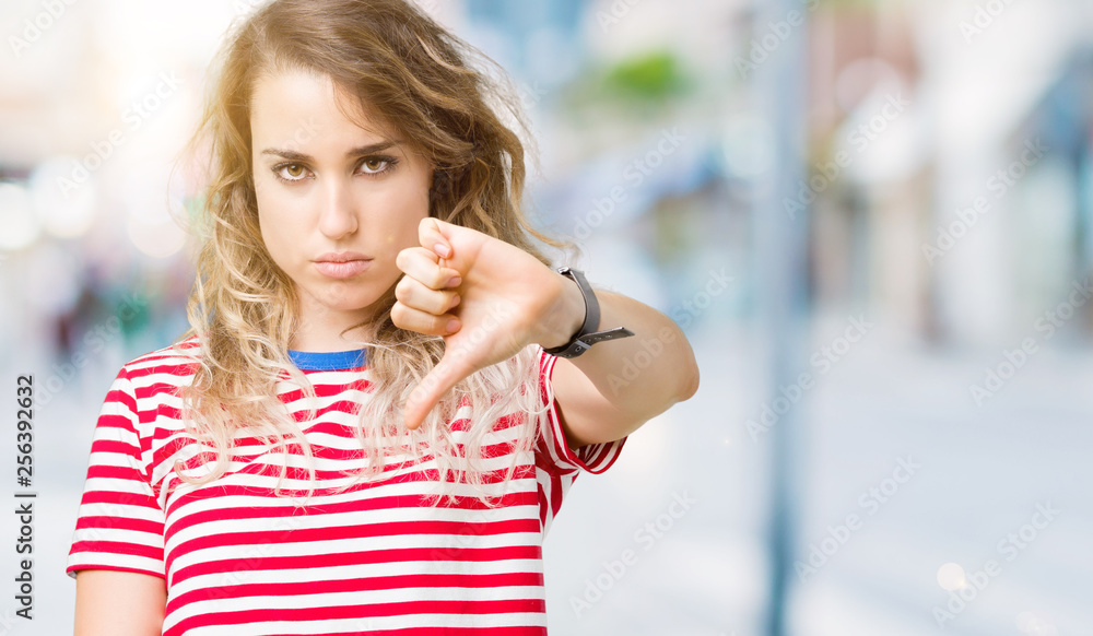 Beautiful young blonde woman over isolated background looking unhappy and angry showing rejection and negative with thumbs down gesture. Bad expression.
