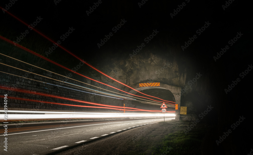 A long exposure photo showing the entrance to the tunnel in mountain with lots of light tails and several traffic signs along the road by night.