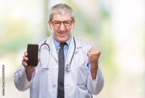 Handsome senior doctor man showing screen of smarpthone over isolated background screaming proud and celebrating victory and success very excited, cheering emotion