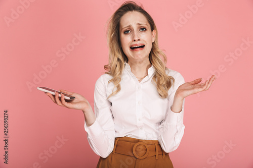Portrait of resentful blond woman 30s in stylish outfit crying while holding and using smartphone
