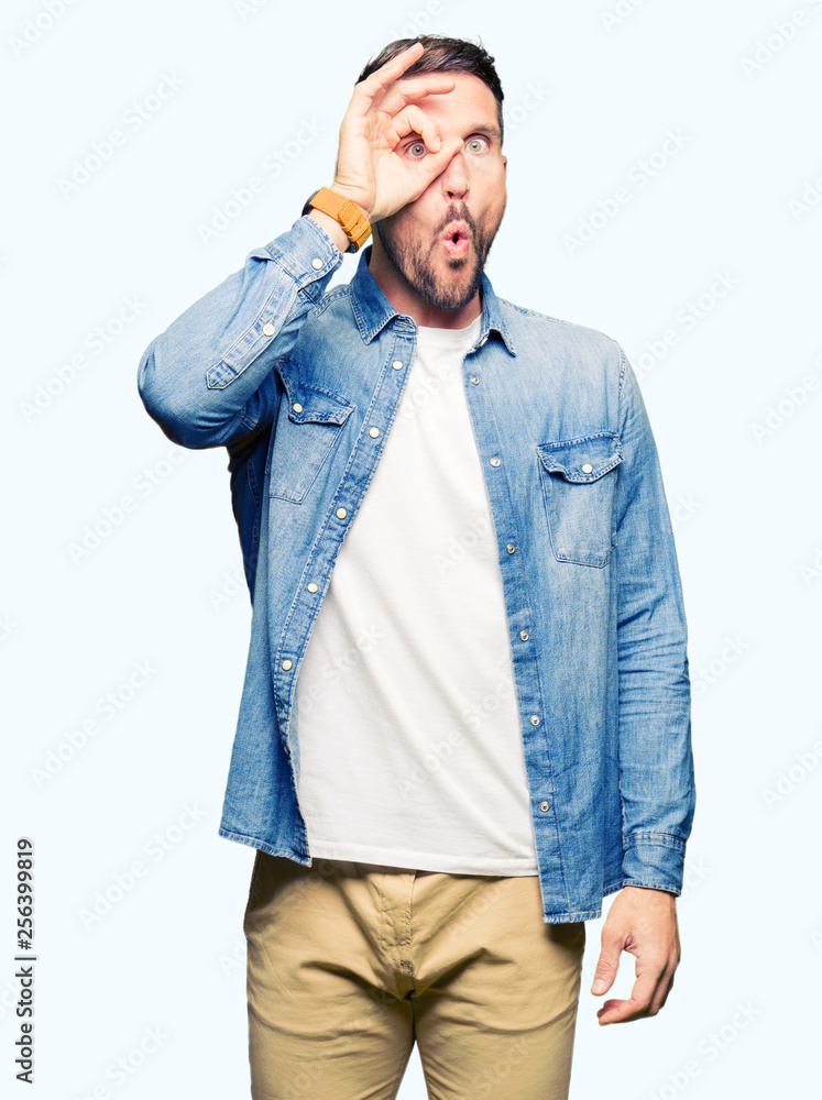 Handsome man with blue eyes and beard wearing denim jacket doing ok gesture shocked with surprised face, eye looking through fingers. Unbelieving expression. Handsome man with blue eyes and beard wearing denim jacket doing ok gesture shocked with surprised face, eye looking through fingers. Unbelieving expression.