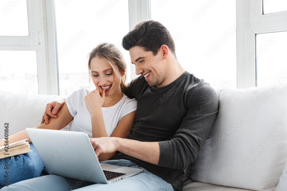 Photo of modern couple using silver laptop while sitting on couch in bright room at home Photo of modern couple using silver laptop while sitting on couch in bright room at home