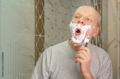 Photo of a man with light gray short hair, shaving his face with a razor and gel or cream in the bathroom closeup
