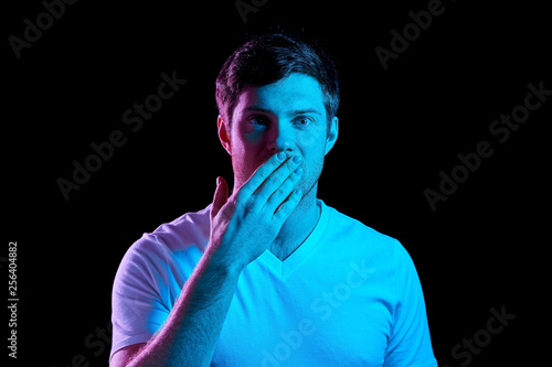 emotion, expression and people concept - portrait of young man in t-shirt covering his mouth by hand over ultra violet neon lights in dark room photo