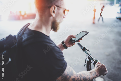 Bearded muscular tattooed hipster in sunglasses using smartphone after riding by electric scooter in the city. Innovative transport