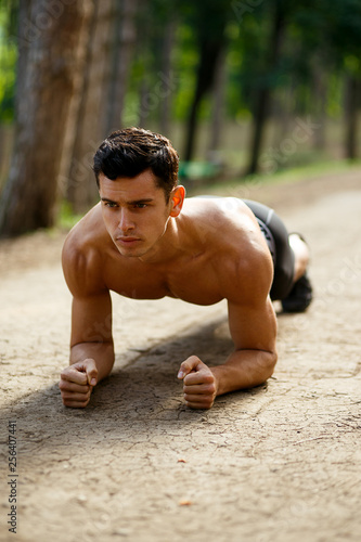 A sportsman doing an abs, an athletic, muscular body, sitting in elbows, isolated on nature background. Healthy lifestyle concept. Park, summer time. Vertical view. © Dragosh