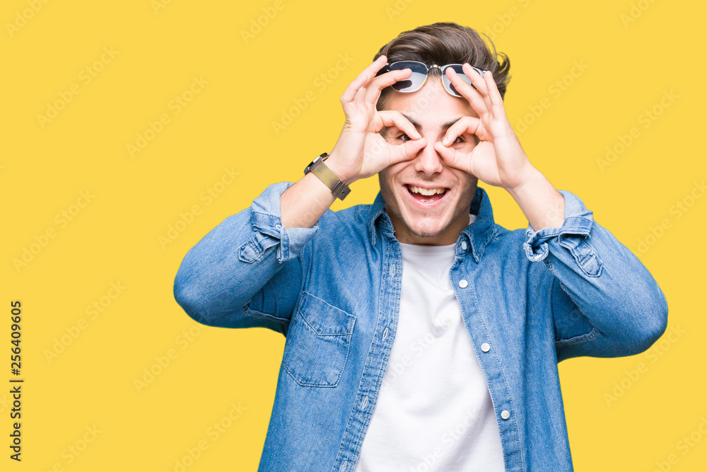 Young handsome man wearing sunglasses over isolated background doing ok gesture like binoculars sticking tongue out, eyes looking through fingers. Crazy expression. Young handsome man wearing sunglasses over isolated background doing ok gesture like binoculars sticking tongue out, eyes looking through fingers. Crazy expression.
