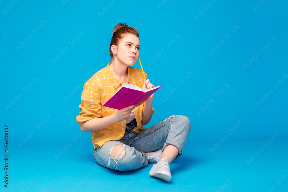 education, high school, inspiration and people concept - red haired teenage girl in checkered shirt and torn jeans with diary or notebook and pencil thinking over grey background - obrazy, fototapety, plakaty 