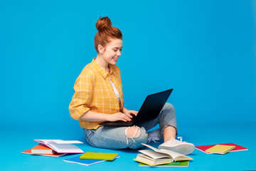 education, high school, technology and people concept - red haired teenage student girl in checkered shirt and torn jeans with books using laptop computer over bright blue background
