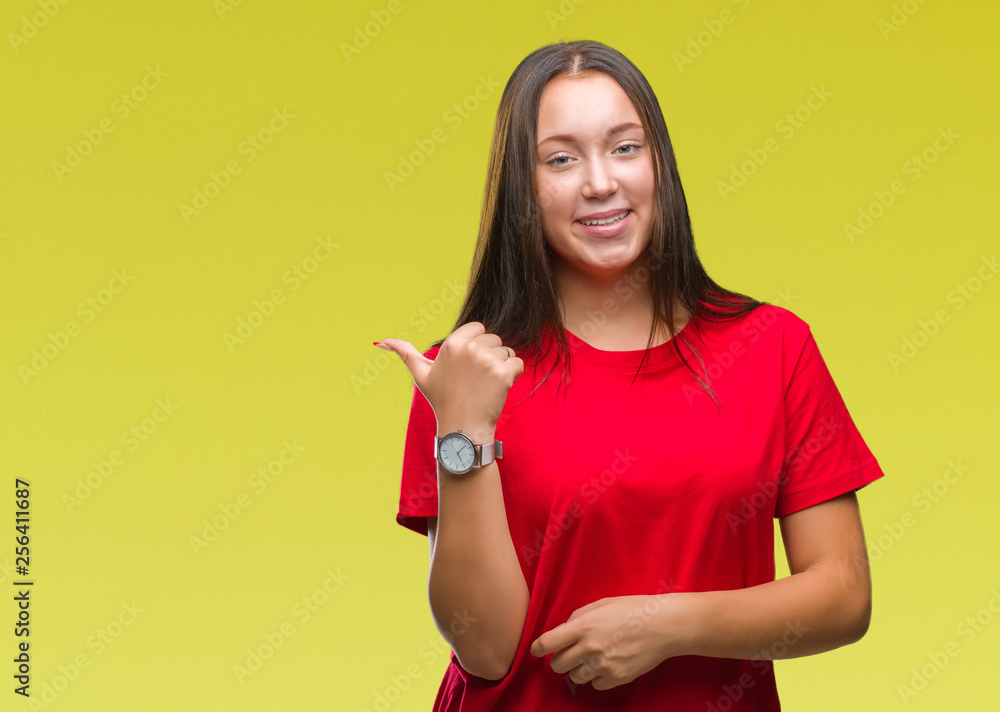 Young beautiful caucasian woman over isolated background smiling with happy face looking and pointing to the side with thumb up. Young beautiful caucasian woman over isolated background smiling with happy face looking and pointing to the side with thumb up.