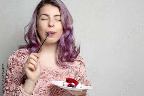 Joyful young girl with purple hair eating tasty cheese cake over grey background. Empty space
