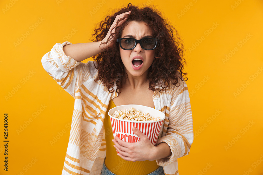 Cute beautiful young curly girl posing isolated over yellow wall background wearing glasses eat popcorn watch film.