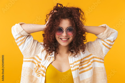 Beautiful young curly girl posing isolated over yellow wall background wearing sunglasses.