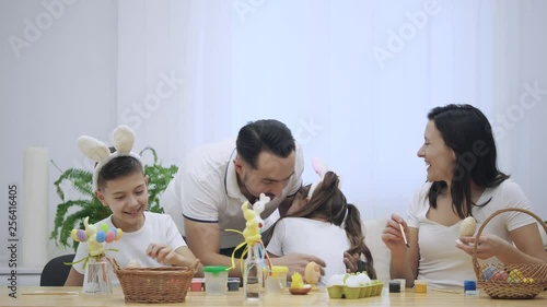 Wallpaper Mural Father is hugging his kids: daughter and son, who are wearing bunny ears, sitting at the holiday table full of Easter decorations. Family members are smiling simultaneously. Slow Motion. Torontodigital.ca