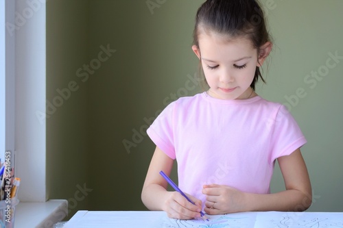 Beautiful caucasian white girl enthusiastically paints a picture with colored pencils. Smiling cute girl in pink T-shirt drawing a picture with pencils on blurred neutral background. Kid learning art