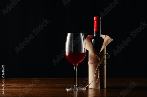 Wine bottle in paper wrapper and glass on wooden surface isolated on black