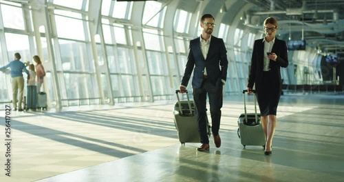 Caucasian business couple of the co-workers travelling on the working trip, walking the airport with suitcases on wheels and talking.