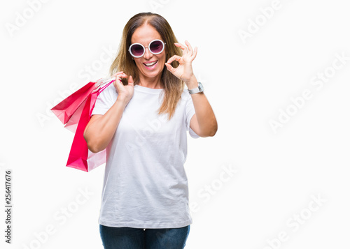 Middle age hispanic woman holding shopping bags on sales over isolated background doing ok sign with fingers, excellent symbol
