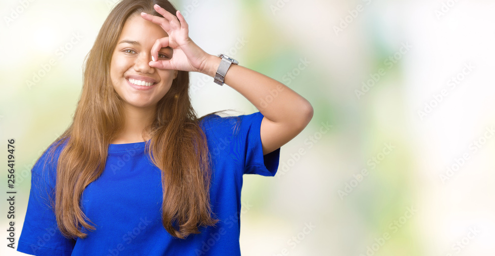 Young beautiful brunette woman wearing blue t-shirt over isolated background doing ok gesture with hand smiling, eye looking through fingers with happy face.