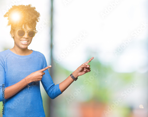 Beautiful young african american woman wearing sunglasses over isolated background smiling and looking at the camera pointing with two hands and fingers to the side.
