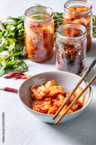 Fotografie Korean traditional fermented appetizer kimchi cabbage and radish salad, hot spicy anchovies fish snack served in glass jars and bowl with Vietnamese oregano and chili peppers over grey blue table
