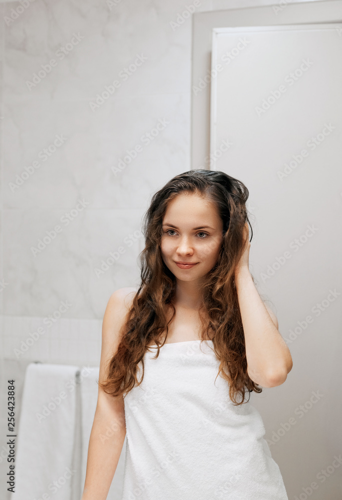 Beautiful young woman touching her wet hair with hands and smiling..Female looking of the mirror in bathroom. Care hair and skin. Girl uses protection moisturizing cream. Beautiful young woman touching her wet hair with hands and smiling..Female looking of the mirror in bathroom. Care hair and skin. Girl uses protection moisturizing cream.