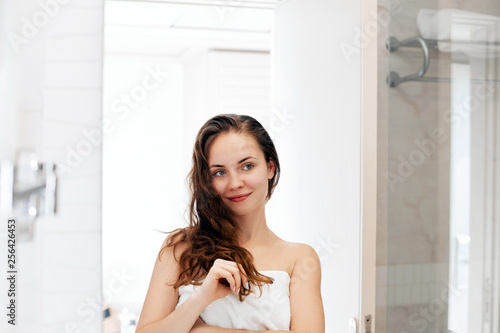 Hair and body care. Woman touching hair and smiling while looking in the mirror.Portrait of happy girl with wet hair in bathroom applying conditioner and oil. Girl uses protection moisturizing cream