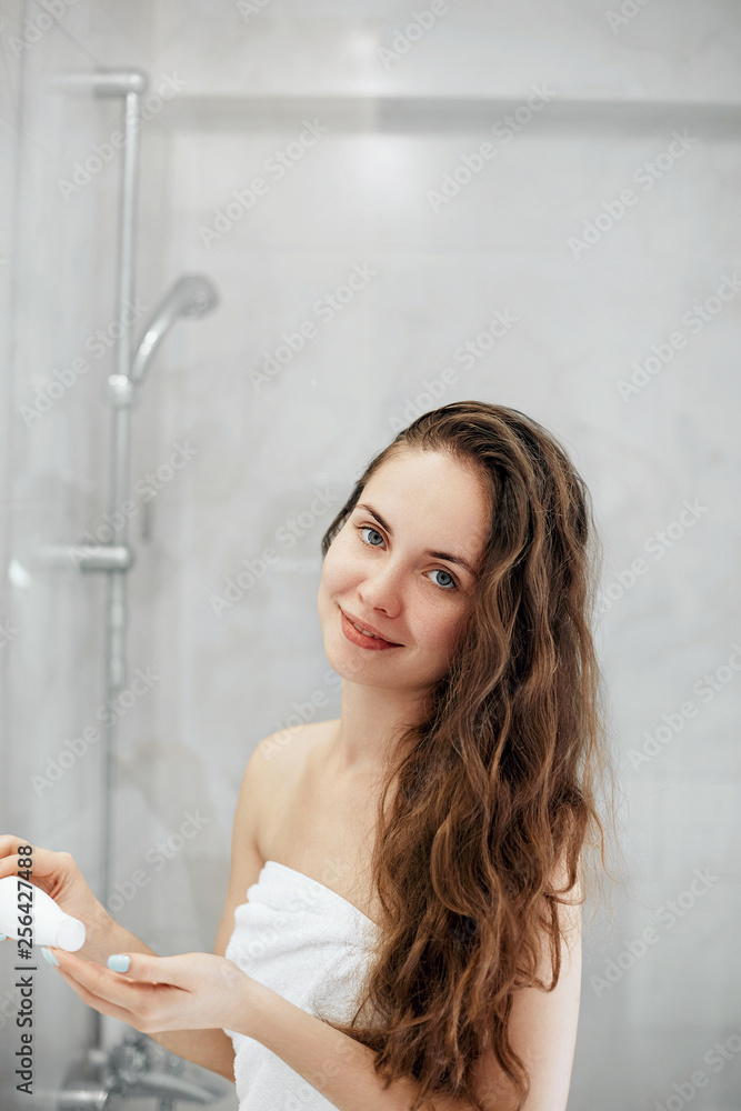 Beautiful young woman applying lotion for hair and smiling while standing in front of the mirror in bathroom. Care hair and skin. Girl uses protection moisturizing cream. Beautiful young woman applying lotion for hair and smiling while standing in front of the mirror in bathroom. Care hair and skin. Girl uses protection moisturizing cream.