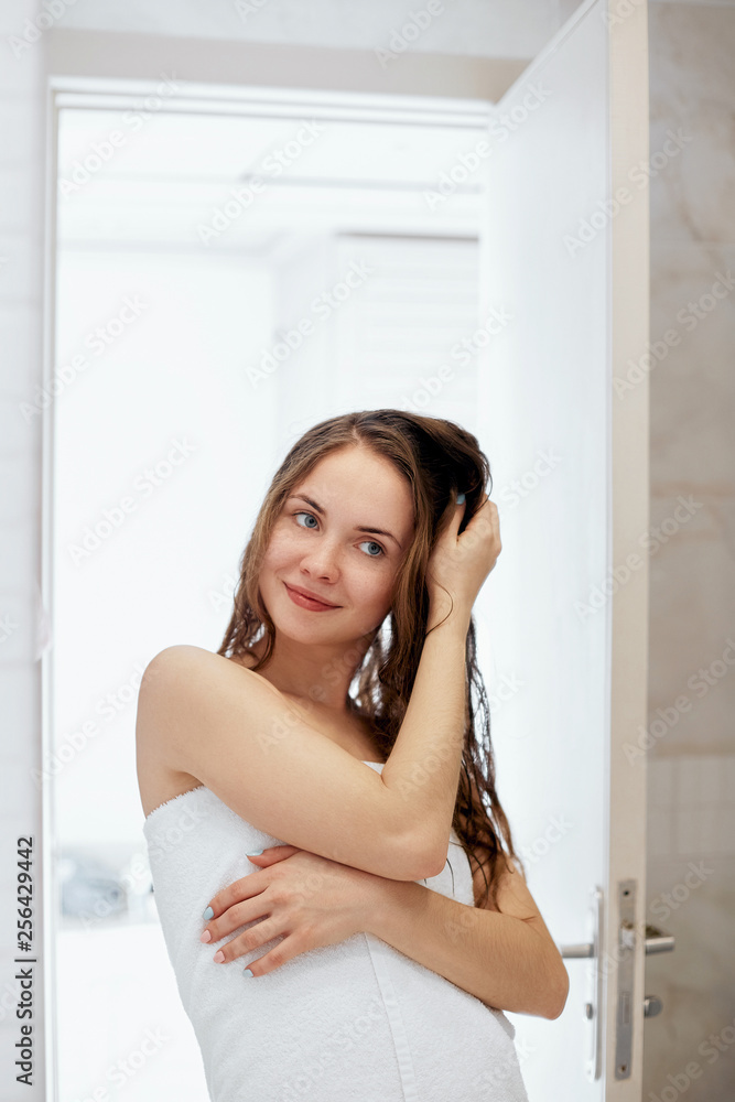 Hair and body care.Beautiful young woman touching her wet hair with hands and smiling while standing in front of the mirror in bathroom. Girl uses protection moisturizing cream. Hair and body care.Beautiful young woman touching her wet hair with hands and smiling while standing in front of the mirror in bathroom. Girl uses protection moisturizing cream.